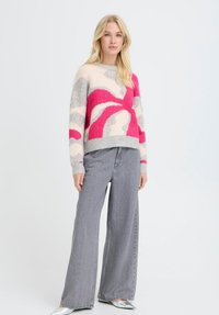 Blonde Frau trägt einen pullover mit abstraktem Muster in Rosa, Grau und Creme, weite graue Jeans und silberne spitze Schuhe und steht.