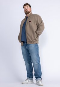 Svetle hnědá zipová bunda s texturovaným designem, pod ní modré tričko a světlé modré džíny. Doplněno bílými teniskami.