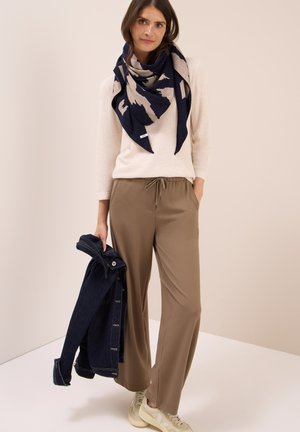 Maglione beige, pantaloni verde oliva con cordino, sciarpa a fantasia blu navy, giacca di jeans scura in mano e sneakers chiare. Sfondo neutro.