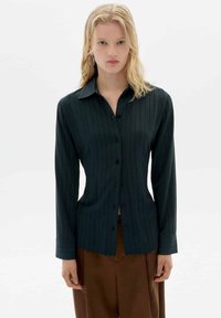 Blonde Frau in einem dunkelblauen gestreiften Hemd mit Knopfleiste und braunen Hosen, die vor einem hellen einfarbigen Hintergrund steht.