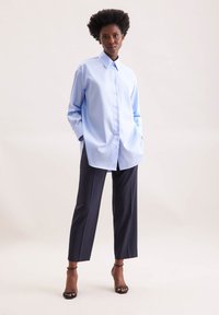 Camicia oversize azzurro chiaro con colletto a punta, abbinata a pantaloni sartoriali blu scuro e sandali neri con tacco. Tessuto dalla superficie liscia.