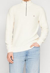 Muž oblečený v texturovaném bílém svetru na zip s půlkou, v kombinaci se světle modrými džíny, s jednou rukou v kapse a druhou volně položenou po boku.