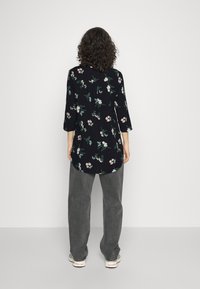 Person med lockigt hår som bär en svart blommig långärmad topp, grå jeans och vita sneakers, stående mot en vit slät vägg.