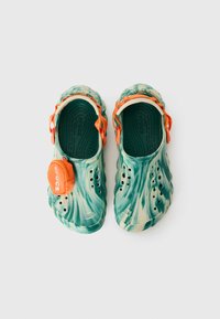 Des Crocs marbrés verts et beiges avec des sangles orange et une petite pochette zippée orange attachée sur un fond blanc.