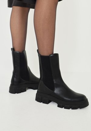 Stivali Chelsea in pelle nera con pannelli elastici laterali, suola in gomma spessa e una consistenza liscia. Progettati per una vestibilità alla caviglia.