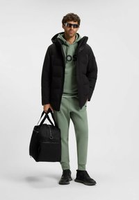 Giacca a vento nera, felpa verde con logo, pantaloni sweat leggeri verdi, borsa da palestra nera e scarpe da ginnastica scure. Occhiali da sole riflettenti completano il look.