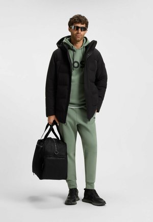 Schwarze Pufferjacke, grüne Kapuzenpulli mit Logo, hellgrüne Jogginghose, schwarze Sporttasche und dunkle Sportschuhe. Reflektierende Sonnenbrille vervollständigen den Look.