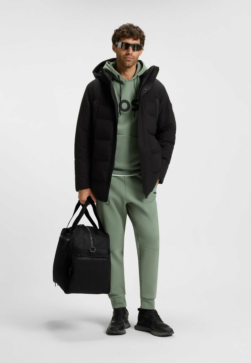 Giacca a vento nera, felpa verde con logo, pantaloni sweat leggeri verdi, borsa da palestra nera e scarpe da ginnastica scure. Occhiali da sole riflettenti completano il look.