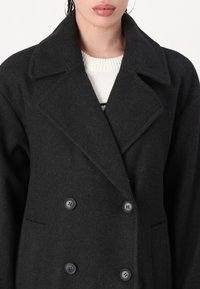 Cappotto doppiopetto in lana nera con ampie revers e due bottoni scuri. Tessuto morbido, vestibilità sartoriale, dotato di tasche laterali.