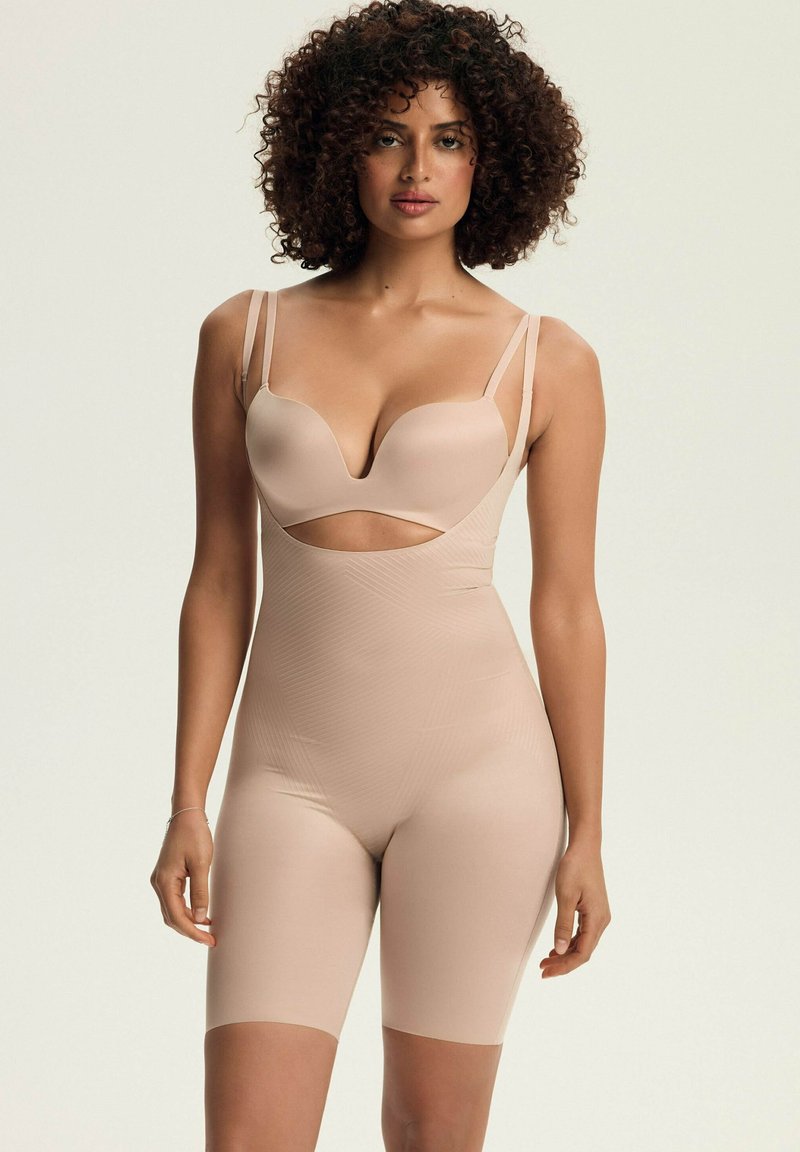 Beżowy bodysuit modelujący o wyciętym designie, z regulowanymi cienkimi ramiączkami i gładką fakturą. Długość do kolan, z dopasowaną sylwetką.