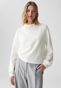 Maglione bianco e soffice con collo alto e maniche lunghe, abbinato a pantaloni grigi plissettati. Tessuto morbido con una silhouette rilassata.