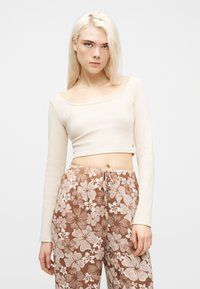 Νεαρή γυναίκα με μακριά ξανθά μαλλιά, ντυμένη με κρεμ μακρυμάνικο κοντό τοπ και καφέ φλοράλ παντελόνια με κορδόνι στη μέση, στέκεται μπροστά σε απλό φόντο.