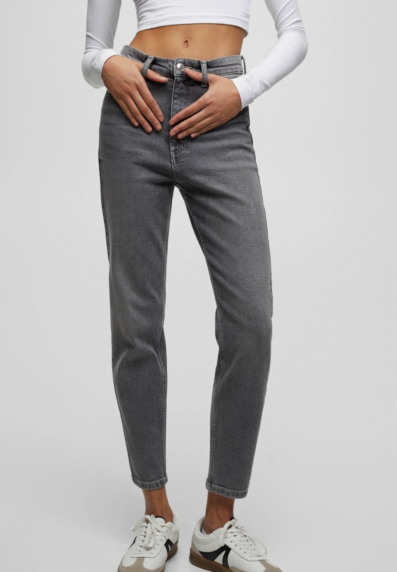 Femme portant un jean gris taille haute, des baskets blanches et un crop top blanc à manches longues, mains reposant près de la ceinture.