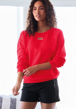 Frau mit lockigem Haar, die ein knallrotes Sweatshirt und schwarze kurze Hosen trägt und in weichem natürlichem Licht drinnen steht.