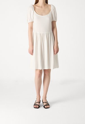 Biała sukienka z bufiastymi rękawami, kwadratowym dekoltem, marszczonym wcięciem w talii i zwiewnym dołem. Połączona z czarnymi sandałami na cienkich paskach i płaską podeszwą.