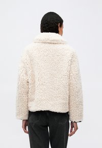 Persoană purtând o jachetă scurtă, de culoare crem, din fleece texturat, cu un guler larg și pantaloni negri, văzută din spate, pe un fundal simplu.