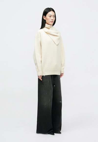 Prevelik krem pulover z teksturiranim, obrnjenim ovratnikom; v kombinaciji z črnimi širokimi hlačami s madeži barve in črnimi konicami čevljev.
