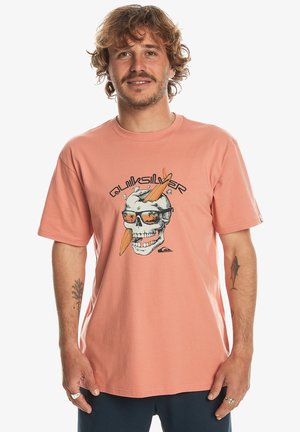 Short-sleeve, coral T-shirt made of cotton. Features a graphic of a skull with sunglasses and surfboards, with "Quiksilver" text above.