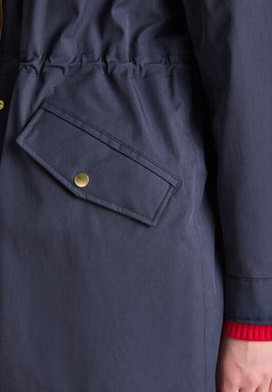 Vrecko námorníckej modrej bundy s jedným zlatým cvokom, elastickým pásom hore a červeným rukávom svetra viditeľným na zápästí.