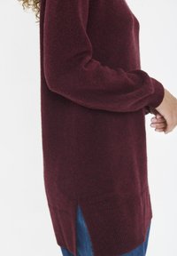 Vrouw in een bordeauxrode wollen trui met een zijspleet over blauwe jeans, weergegeven van halverwege de romp tot de heupen tegen een witte achtergrond.