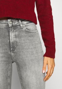 Suéter rojo de punto con puños acanalados, combinado con unos pantalones vaqueros de denim gris de tiro alto que cuentan con cinco bolsillos y un cierre de botón en la parte delantera.