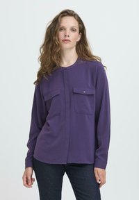 Donna che indossa una camicia bottonata viola a maniche lunghe con due taschini sul petto, in piedi contro uno sfondo chiaro e uniforme.