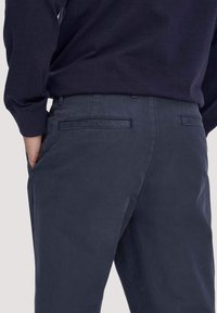 Mann trägt marineblaue Hose mit hinteren Leistentaschen und passendem marineblauen Langarmhemd, Hand in Seitentasche, leicht abgewandt.