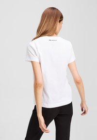 Weißes Baumwoll-T-Shirt mit kurzen Ärmeln, rundem Halsausschnitt und einem dezenten Logo auf der Rückseite. Kombiniert mit schwarzen Hosen.