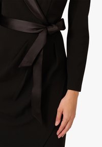 Schwarzes Kleid mit glatter Textur, ausgestattet mit einem gebundenen Satin-Gürtel in der Taille und langen Ärmeln. Das Design ist figurbetont mit drapierten Stoffdetails.
