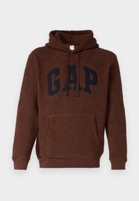 Hnědá fleecová mikina s kapucí a klokáním kapsou, černé logo "GAP" na přední straně a žebrované manžety a lem. Měkká, strukturovaná tkanina.