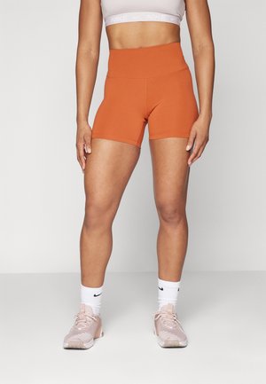 Žena v oranžových atletických šortkách s vysokým pasem, bílém sportovním topu, bílých ponožkách Nike a růžových běžeckých botách, stojící na jednobarevném světlém pozadí.
