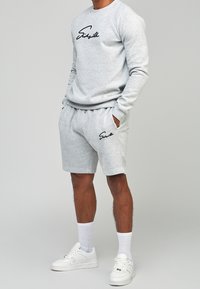 Felpa grigia con logo nero sul petto, abbinata a pantaloncini grigi coordinati. Il modello indossa sneakers bianche e calzini al polpaccio.