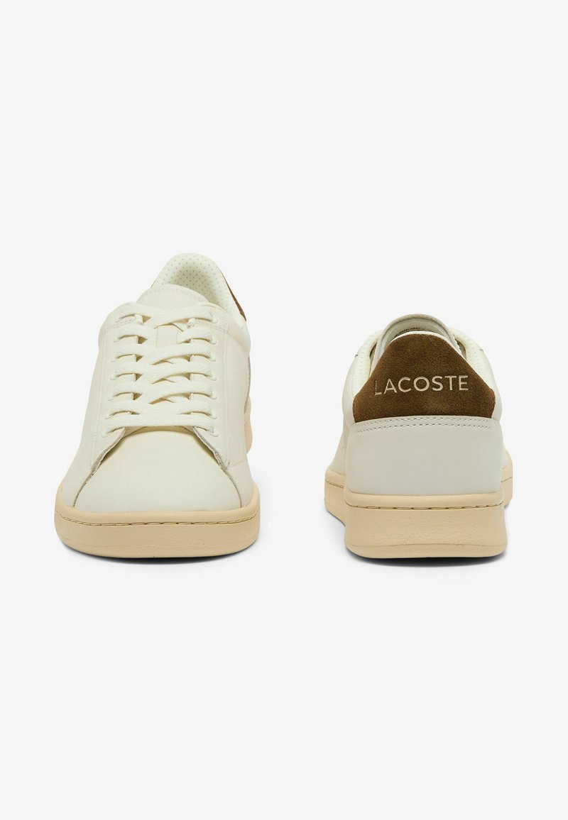 Coppia di sneakers Lacoste color avorio con suola beige, che mostra la vista frontale della scarpa sinistra e la vista posteriore della scarpa destra con linguette marroni sul tallone.