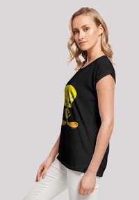 Svart t-skjorte med et stort, fargerikt bilde av Tweety Bird i gult. Korte ermer og en avslappet passform, iført med hvite bukser.
