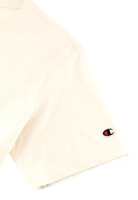 Beige Baumwoll-T-Shirt mit Rundhalsausschnitt, kurzen Ärmeln und einem Champion-Logo-Patch mit roten und schwarzen Akzenten nahe dem linken Ärmel.