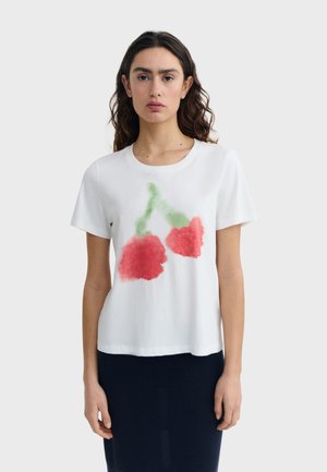 Femme aux cheveux ondulés et foncés portant un t-shirt blanc à manches courtes avec un imprimé floral abstrait rouge et vert sur le devant, assorti à une jupe foncée.