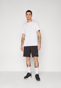 Witte T-shirt met korte mouwen, zwarte sportshorts, zwarte sneakers met groene accenten, witte sokken. Tatoeages zichtbaar op de armen. Eenvoudige achtergrond.
