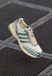 Sneaker Adidas bianco e verde con tomaia in rete e suola beige sospesa su uno sfondo di grata metallica.