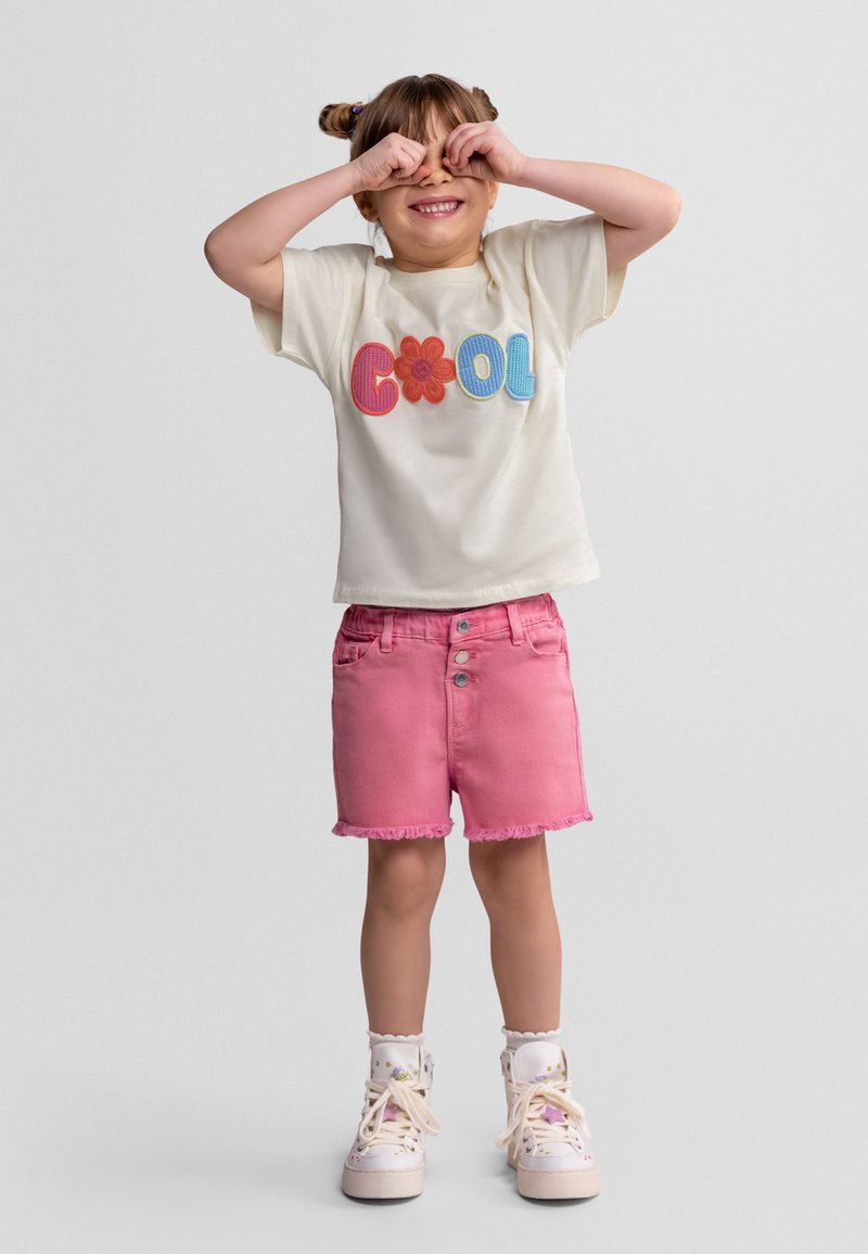 Jeune fille souriante se couvrant les yeux avec les mains, portant un t-shirt blanc "COOL", un short rose et des baskets blanches avec des chaussettes.