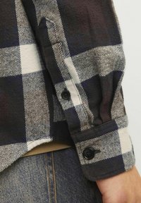 Brun og navy plaid flannel skjorte med sorte knapper. Materialet er blødt og tekstureret; har en rund nederdel og manchetter med synlige syninger.