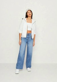 Giacca bianca con zip indossata sopra un crop top aderente, abbinata a jeans blu a gamba larga e sneakers bianche, completata da un berretto leggero.