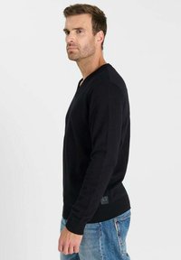 Zwarte v-hals trui van zacht materiaal, met geribbelde details op de schouders en manchetten, gecombineerd met lichtblauwe spijkerbroek.