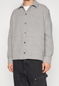 Camicia grigia con bottoni, caratterizzata da un motivo testurizzato, con colletto e bottoni neri, abbinata a pantaloni scuri.