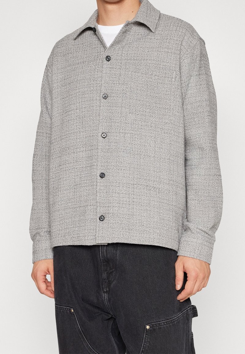 Camicia grigia con bottoni, caratterizzata da un motivo testurizzato, con colletto e bottoni neri, abbinata a pantaloni scuri.