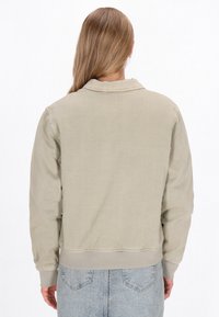 Világos khaki színű bomber dzseki, gallérral, bordás mandzsettákkal és aljjal, texturált anyagból készült. A hátsó nézet egyszerű dizájnt mutat, látható záródás nélkül.