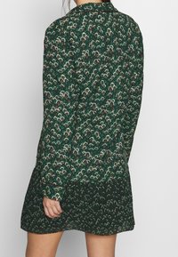 Abito verde a fantasia con maniche lunghe, caratterizzato da un motivo floreale in rosso e bianco. Il tessuto sembra leggero e strutturato, con un orlo leggermente arricciato.