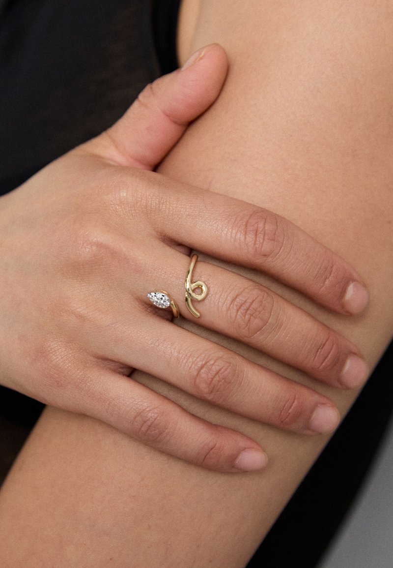 Arany gyűrű, amelyben egy marquise csiszolású kő található, és ívelt formája van, egy kézen viselve, amely egy meszelt karon nyugszik. Sima textura, minimalista stílus.