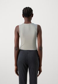 Lys grå ermeløs crop-top laget av glatt stoff, med rund hals og tettsittende design. Brukt sammen med bukser i mørkegrå med vertikale striper.