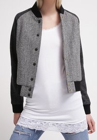 Szary bomber z wzorem w jodełkę, z czarnym ściągaczem na kołnierzu i mankietach. Zawiera napy oraz luźny krój, noszony na białej bluzce z koronkowym wykończeniem.