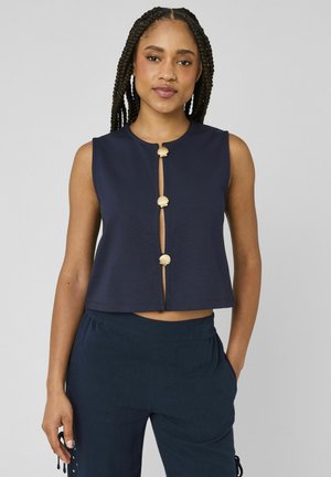 Femme aux cheveux tressés portant un haut sans manches bleu marine avec trois boutons dorés en forme de coquillage et un pantalon assorti bleu marine, debout, la main dans la poche.